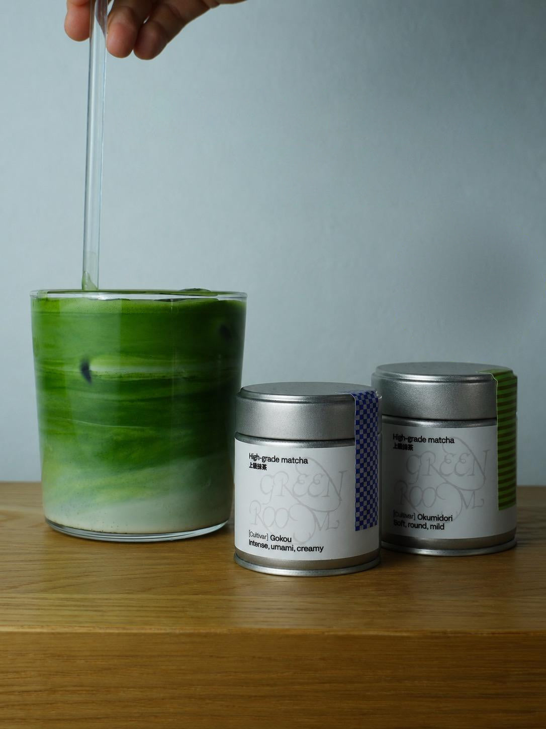 Okumidori and Gokou 30 g tins next to an iced matcha latte being stirred in a glass